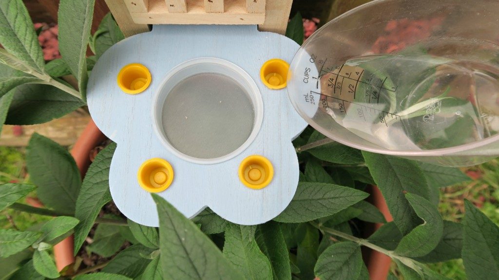 A butterfly-shaped bee watering station with four yellow cups is surrounded by green leaves—perfect to create a butterfly garden. A clear measuring jug, held to the right, pours water into the central reservoir.