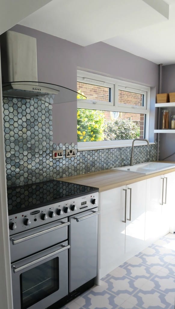 Modern kitchen with white cupboards, hexagonal blue glass mosaic tiles splashback in the Original Style, stainless steel oven and hob, light wood worktop, large sink, and window above the counter letting in natural light.