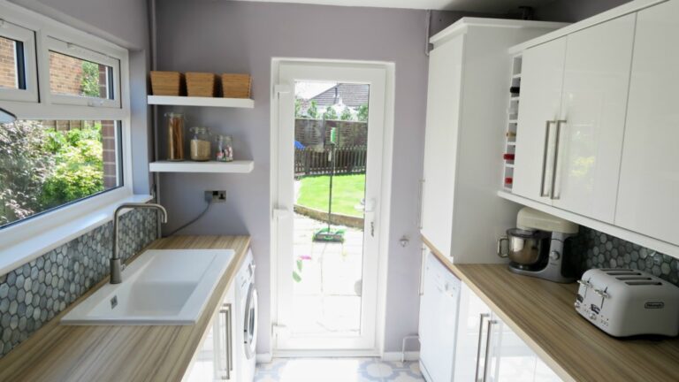 A bright, modern kitchen refurbishment with light wood worktops, white cupboards, a butler sink, appliances, and shelves. A glass door opens to a green garden outside the fence. Sunlight fills this inspiring renovation reveal.