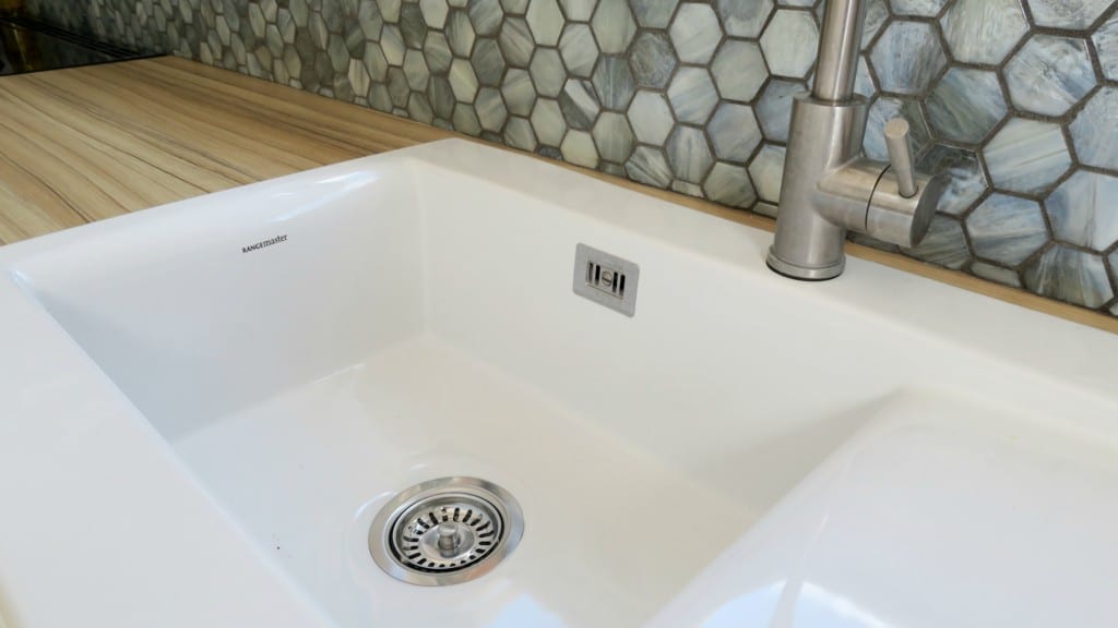A modern white kitchen sink with a silver tap, metal waste, and overflow plate, set against a hexagonal tile splashback and wooden worktop—perfect for your next kitchen renovation or stylish kitchen reveal.