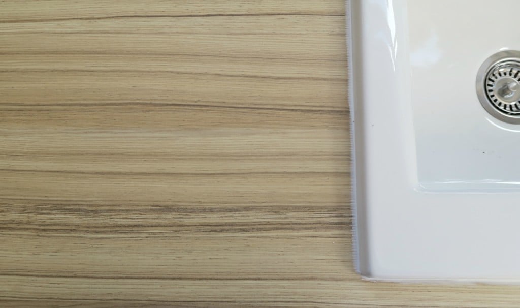 A close-up view of a light wood-grain worktop from a recent kitchen refurbishment, with the edge of a white ceramic sink and a metal waste visible on the right side of the image.
