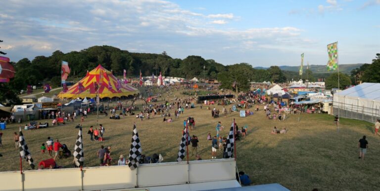 A large outdoor festival scene at Camp Bestival 2016 with crowds of people on a grassy field, colourful tents including a striped circus tent, flags, and various stalls—perfect for a fun-filled day visit under a mostly clear sky with scattered clouds.
