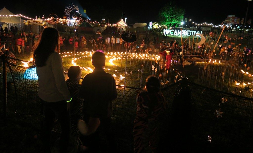 A group of people at night stand behind a fence, enjoying the vibrant festival scene at Camp Bestival 2016, with glowing lights in circular patterns and illuminated decorations. A lit "CAMP BESTIVAL" sign glows in the background.