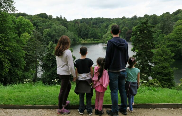 Me and Mine: In August, a family of five stands together on a grassy hill, overlooking a lake surrounded by lush trees beneath an overcast sky, enjoying the scenic view.