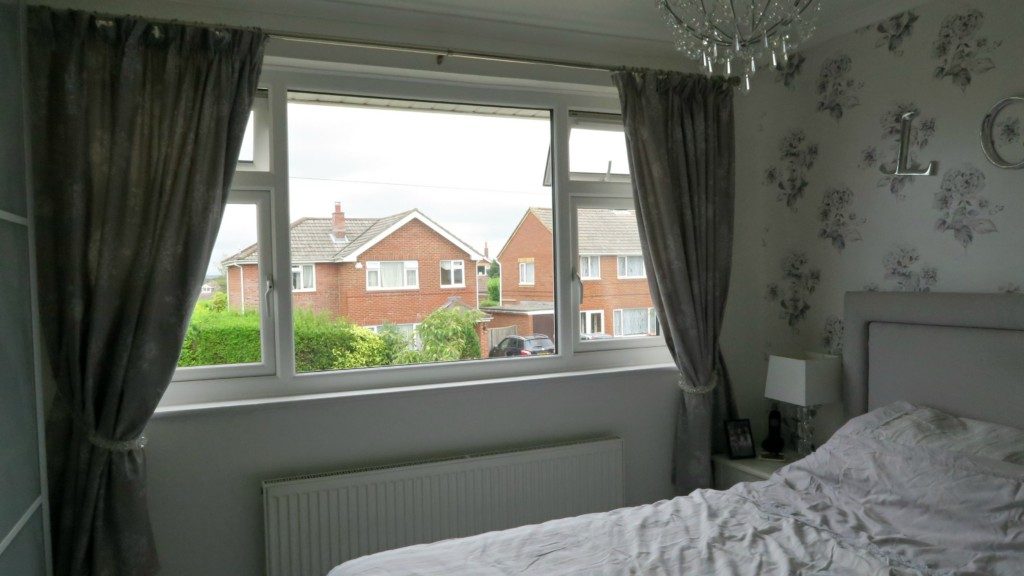 A bedroom with swish grey curtains held by elegant tie-backs, a large window overlooking suburban houses, a white bed, a bedside table with a lamp, floral wallpaper, and a chandelier.