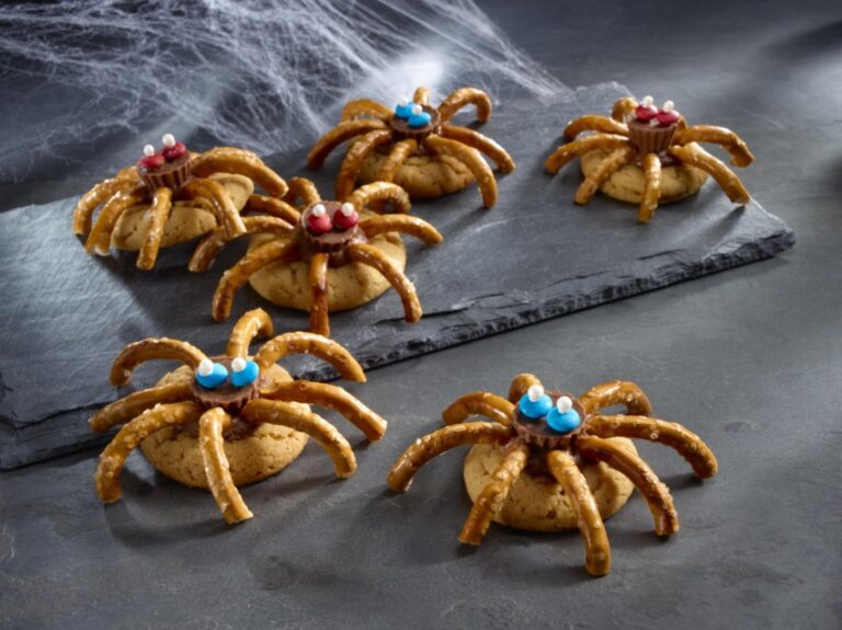 Six spider-themed biscuits with pretzel legs, colourful sweet eyes, and chocolate pieces are arranged on a dark slate tray. Perfect for Halloween recipes, fake cobwebs in the background add to the spooky food and drink display.