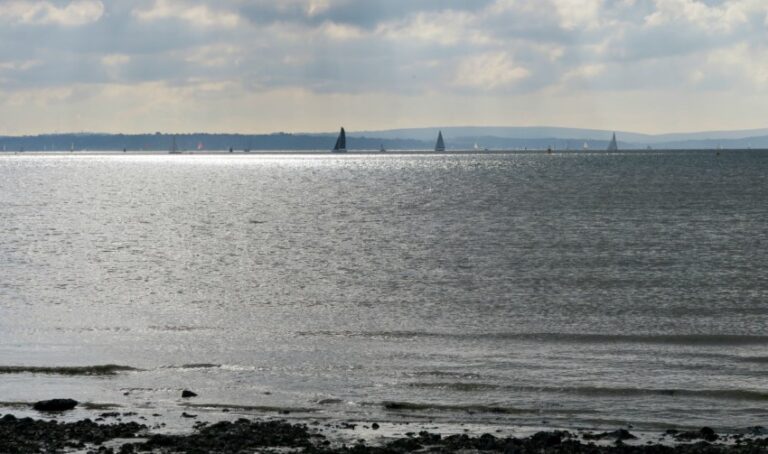 A calm Chilling Beach lies under a cloudy sky, sunlight sparkling on the water. Several sailboats glide in the distance near the horizon, whilst the rocky shoreline in the foreground adds to the tranquil scene.