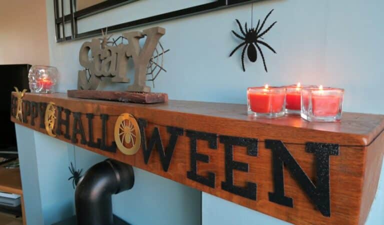A wooden mantelpiece decorated for Hallowe’en with black Happy Hallowe’en letters, fake spiders, red tealight candles, a HomeSense candle holder, and a decorative “Stay” sign against a light blue wall.