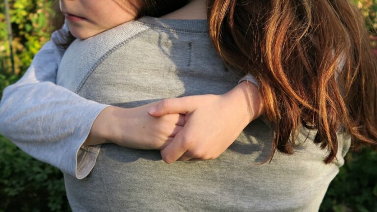 Two kids with long brown hair embrace each other outdoors, wearing grey tops. One child’s face is partially visible, the other’s back is turned. Blurred greenery in the background hints at a bright future ahead.