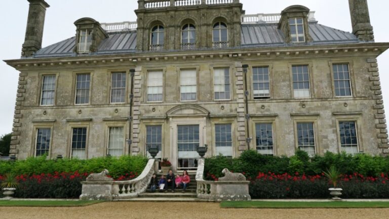 A large, historic stone mansion—Kingston Lacy—is known for its symmetrical windows and two chimneys. Managed by the National Trust, it features a central staircase leading to a garden where people relax among red flowers and greenery.