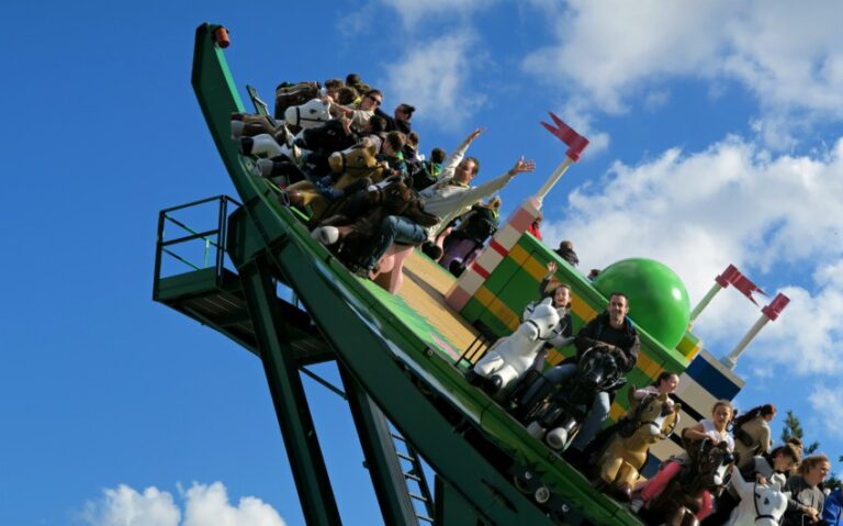 A group of people ride a pirate ship amusement park ride, tilting upwards against a blue sky with scattered clouds. Some riders raise their arms and smile as colourful LEGO-style decorations hint at festive Legoland Fireworks celebrations.