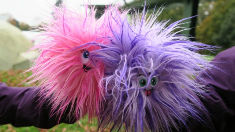 Two fluffy, round puppets—Peeka Puffs—with big eyes and tongues out, one pink and one purple, are held up outdoors. Their long, wild fur adds to their playful, silly charm—ready for a Peeka Puffs Adventure!.