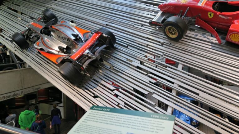 Two Formula 1 racing cars, one silver and one red, are displayed on angled metal tracks in a museum. Visitors stand below, taking in the scene—perhaps fresh from a Beaulieu Firework or Hallowe'en Event nearby. A plaque about the Ferrari F310 is visible in front.