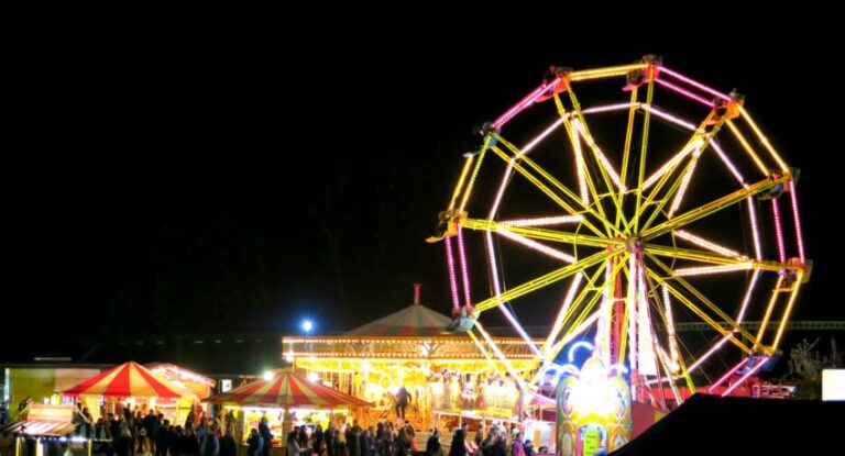 A brightly lit Ferris wheel and carousel glow against the night sky at Beaulieu's lively funfair, where crowds stroll among colourful stalls and attractions during the festive Hallowe'en Event and Beaulieu Firework display.