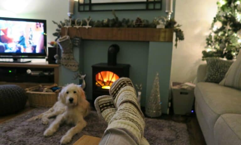 A person wearing cosy, patterned socks relaxes with their feet up near a fireplace; a fluffy dog lies nearby. A TV, holiday decorations, and a lit Christmas tree create the perfect cosy evening atmosphere.