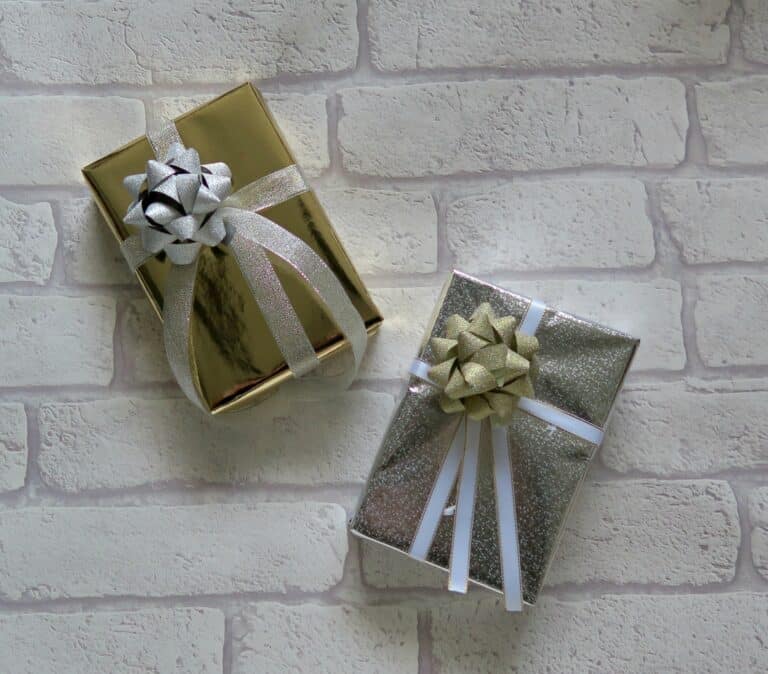 Two gift boxes against a white brick background; one wrapped in gold paper with a silver bow, the other in silver glitter paper with a gold bow—perfect Christmas presents from HomeSense, both decorated with ribbon and priced under £10.