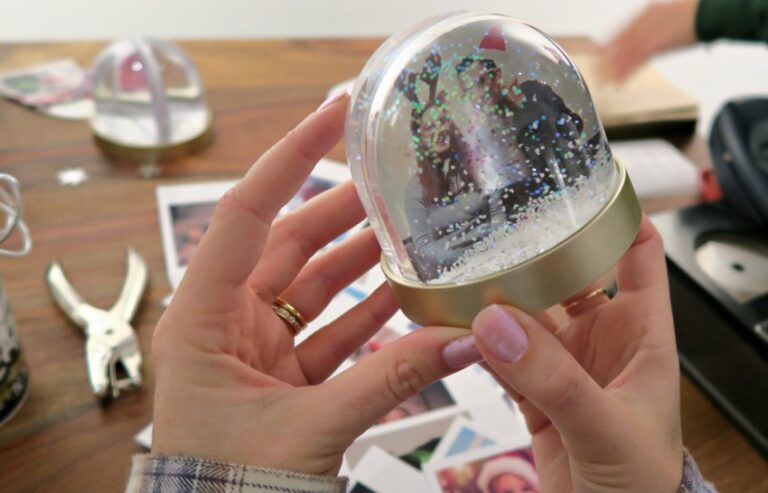 A person holds a small snow globe with a photo of two people inside. Crafting supplies, printed photos from Photobox, and another snow globe rest on a wooden table in the background.