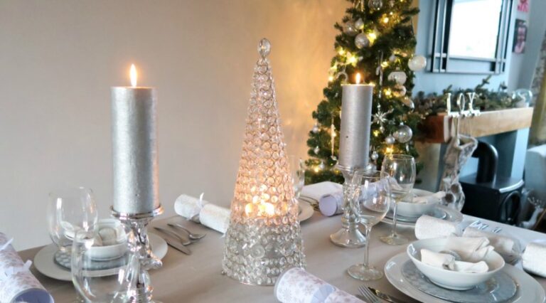 A festive Silver Christmas Table set with silver candles, a decorative glass Christmas tree centrepiece, elegant place settings, and a Christmas tree with white ornaments and lights in the background.