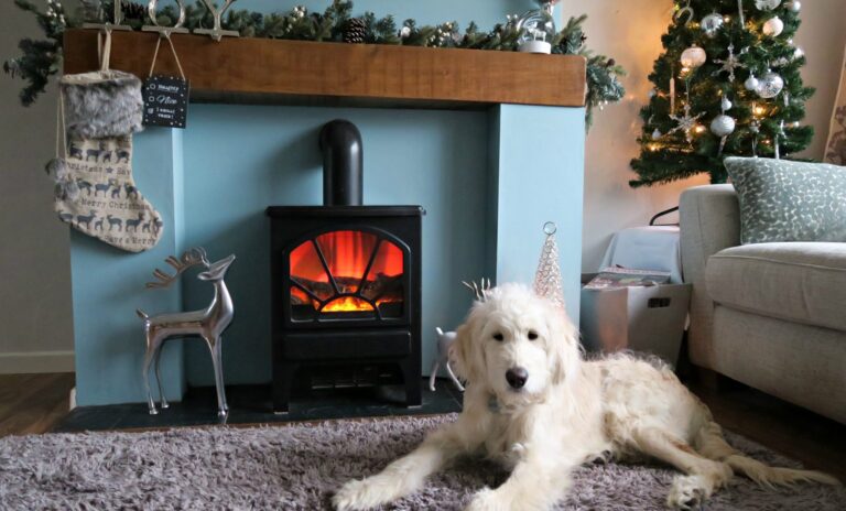 A fluffy white dog lies on a grey rug in front of a lit fireplace decorated for Christmas, with stockings, garlands, and a metal reindeer—creating the perfect cosy spot if you want to work with me during the holidays.