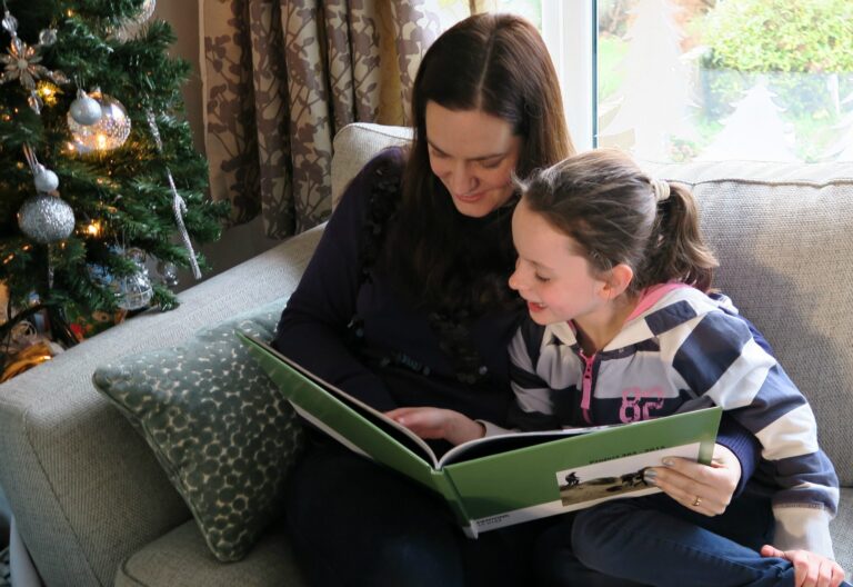 A woman and a young girl sit on a sofa by a Christmas tree, smiling and looking at a photo album from 2016 together. The cosy room, aglow with natural light and festive decor, feels extra special after their recent refurbishment.