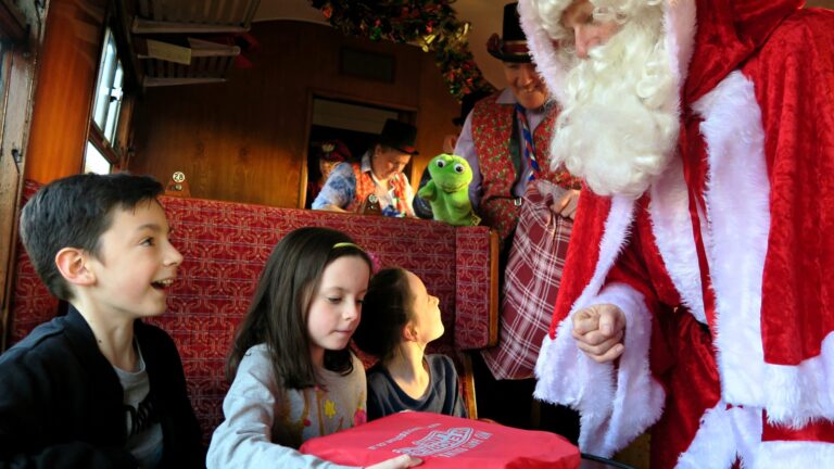 Three children sit in a festive Christmas train carriage on the Watercress Line, excitedly interacting with Father Christmas during the Santa Special as he hands them a present, while entertainers—including one with a puppet—watch and smile in the background.