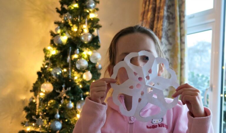 A child in a pink Hello Kitty hoodie holds a paper snowflake in front of their face, showing off their Snowflake Crafts. Behind them, a decorated Christmas tree with white lights and baubles stands near a window with curtains.
