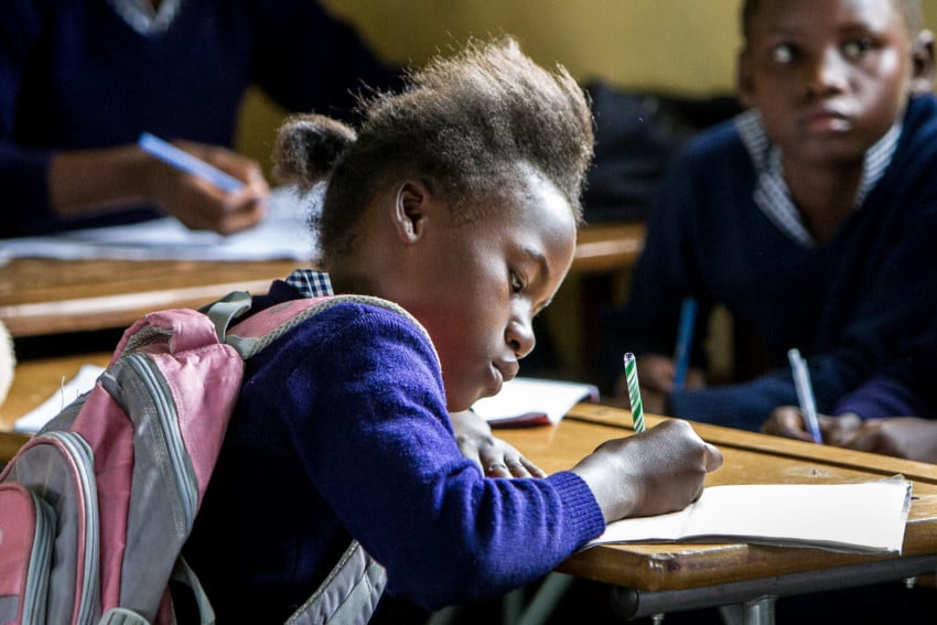 A young girl with a pink rucksack writes in a notebook at her desk in a classroom, whilst another pupil looks on. Both children are focused and engaged—just like when I plan budget meals for £2 each to help feed my family efficiently.