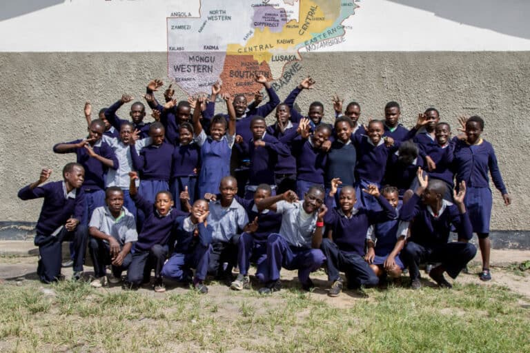 A group of smiling schoolchildren in navy blue uniforms pose energetically outside against a wall painted with a colourful map of African countries, sharing their favourite cheap family meals and ideas to feed family on a budget.