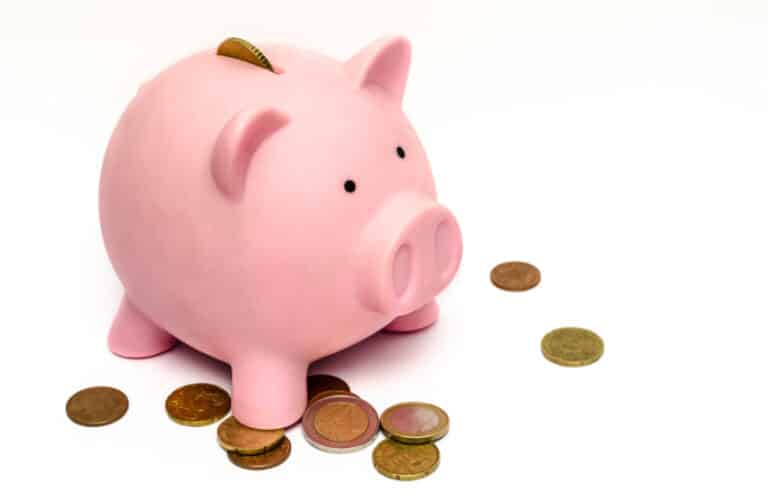 A pink piggy bank with a coin inserted in the slot stands on a white surface, surrounded by scattered coins—perfect for anyone interested in money saving tips.