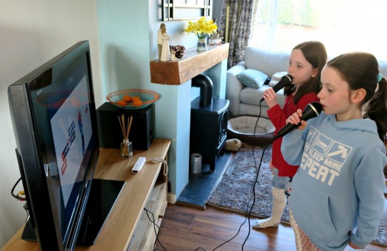 Two young girls sing into microphones while playing The Voice Wii Game on a TV in a cosy living room with wooden floors and a fireplace.