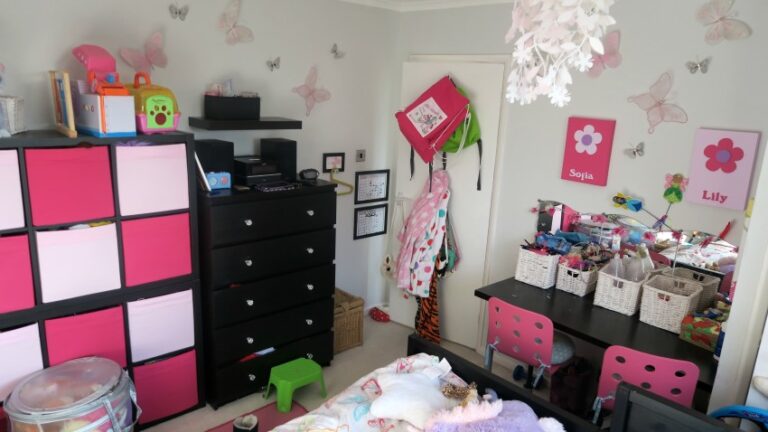 A colourful, organised Twin Girls’ Bedroom with pink and black storage cubes, a black chest of drawers, shared vanity with two pink chairs, baskets, and butterfly decorations on light grey walls. Coats and bags hang on a door.