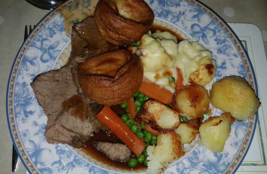 A plate of traditional roast dinner with sliced beef, Yorkshire puddings, roast potatoes, peas, carrots, cauliflower cheese, and brown gravy is served on a floral patterned plate—perfect for weekend comfort or week 8 photos.