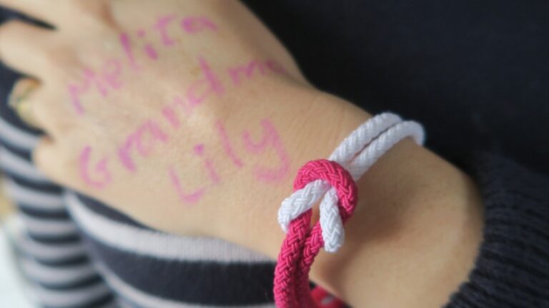 A hand with "Nolita Grandma Lily" written in pink marker, wearing a plaited bracelet with intertwined red, white, and pink cords—a touching nod to cancer awareness—rests gently on dark-striped fabric.
