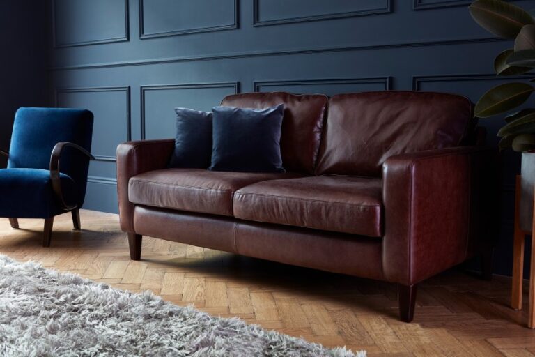 A brown leather sofa with two dark blue cushions sits on a parquet wood floor beside a blue armchair. A light grey shag rug and dark blue panelled wall add a modern twist to the traditional design of the space.
