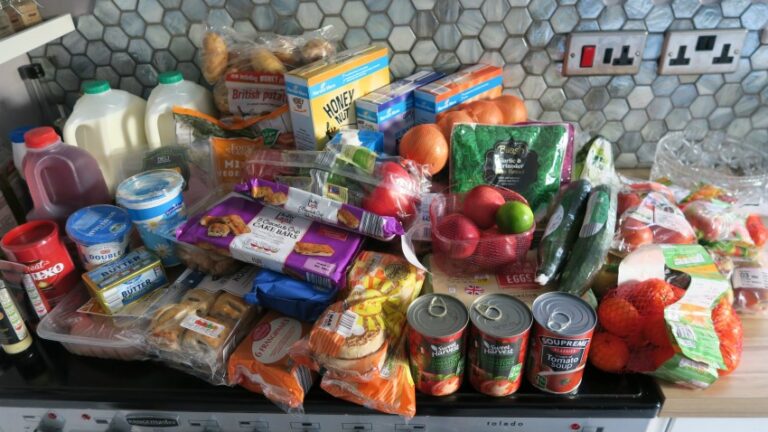 A grocery haul featuring milk, juice, eggs, bread, fruit, vegetables, cheese, yoghurt, cereals, and packaged foods is spread out on a kitchen worktop in front of a tiled splashback—perfect for a weekly meal plan.