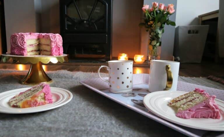 A cosy indoor picnic for two with plates of pink-iced cake, two white mugs on a tray, a whole layered cake on a stand, lit candles, and a vase of pink roses arranged on a carpeted floor—perfect for Valentine’s Day.