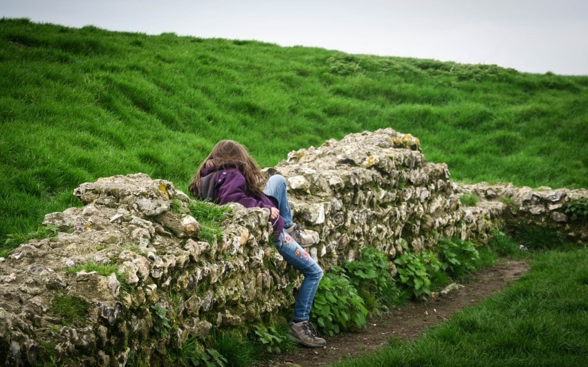 A person with long brown hair, wearing a purple jacket and jeans, sits on an old stone wall surrounded by green grass, their back facing the camera—capturing a quiet moment for this Weekend in Pictures.