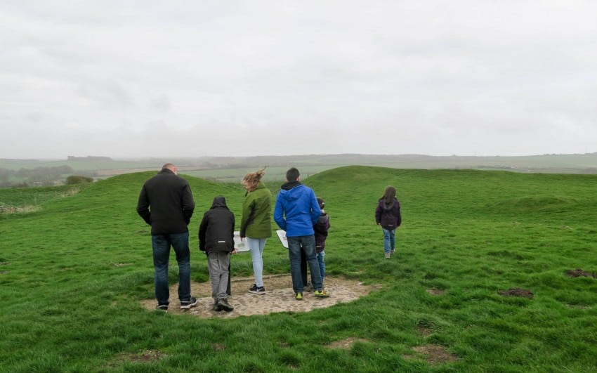 A group of six people, including adults and children, stand on a grassy field under an overcast sky. Captured during Week 15, most are facing away and looking at the landscape, whilst one person walks ahead alone.