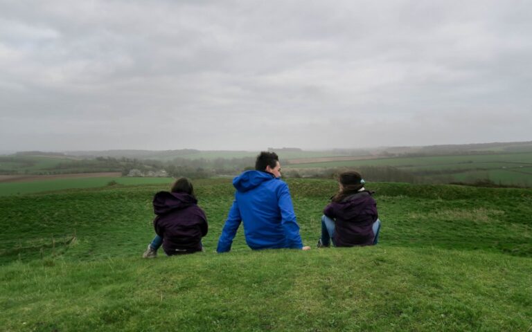 Three people in jackets sit on a grassy hill, facing away and looking out over a wide, green, gently rolling landscape under a cloudy sky—a peaceful moment from our weekend in pictures.