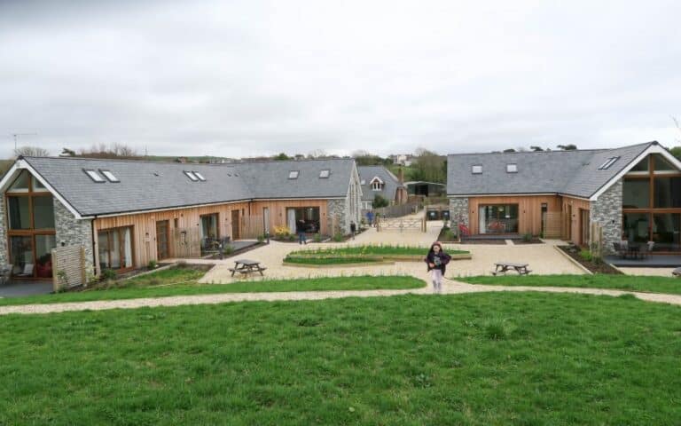 A courtyard with two modern, barn-style buildings facing each other in Upwey Dorset, separated by pathways and flowerbeds. A person stands on the path leading to the courtyard, surrounded by green grass under a cloudy sky—perfect for a Sykes Cottages escape.