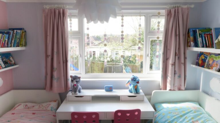 Bright Twin Girls’ Bedroom with two beds, a white desk in between, pink and blue chairs, soft toys on the desk, shelves of books, pink curtains, and a large window letting in natural light. Perfect for your next Bedroom Reveal!.