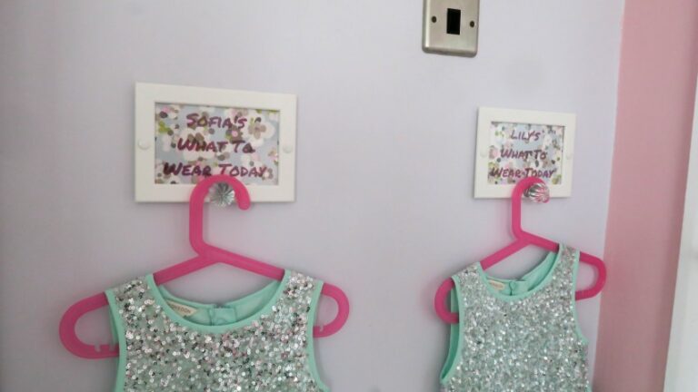 Two sparkly, mint-coloured dresses on pink hangers hang in a Twin Girls’ Bedroom under signs labelled “Sofia’s What to Wear Today” and “Lily’s What to Wear Today,” with a light background and a pink border on the right.