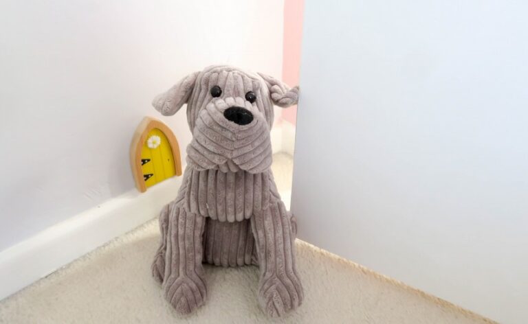 A plush, corduroy dog toy sits on a white carpeted floor near the edge of a white wall in a charming Twin Girls' Bedroom. Behind it, a small yellow door with two black stars is attached to the wall.