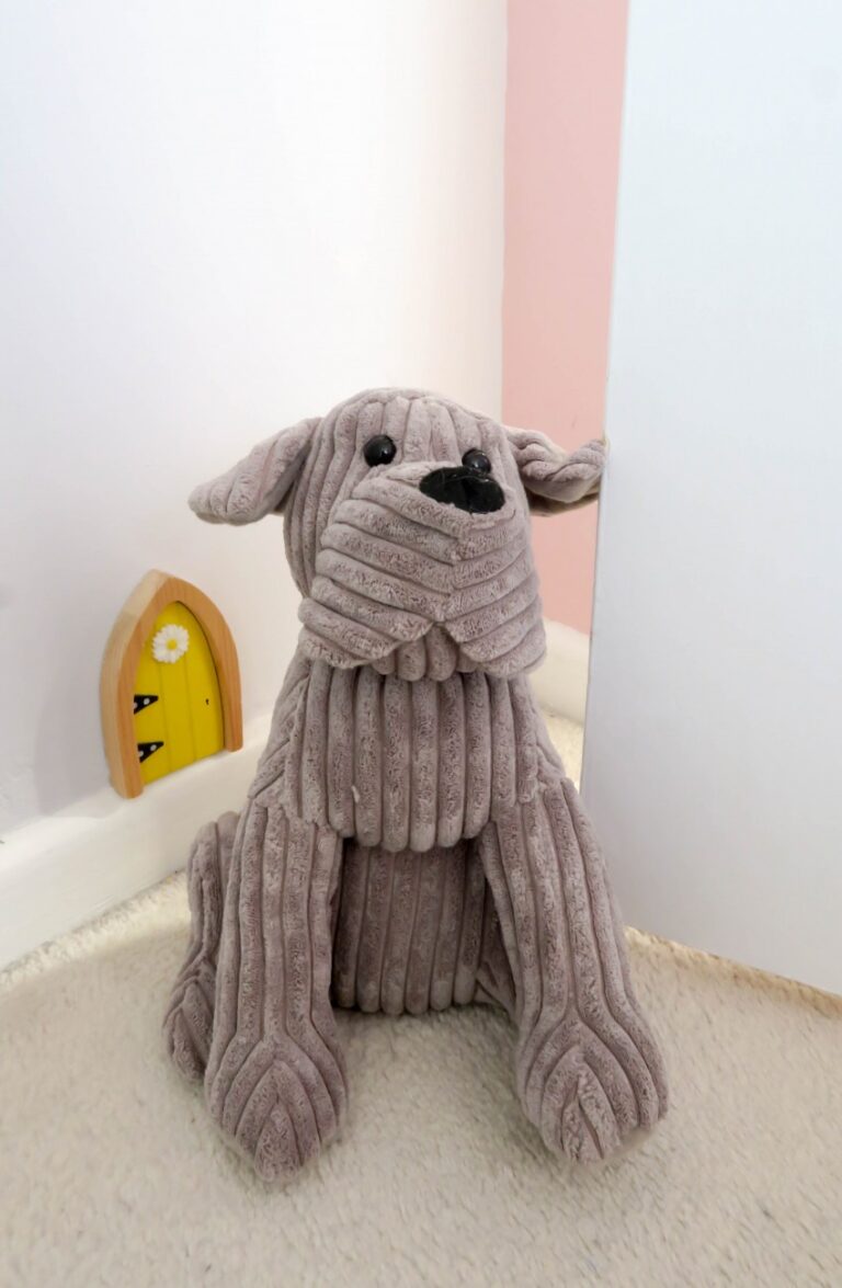 A soft, ribbed grey stuffed dog sits on a light carpet next to a white wall and a small yellow door with a heart-shaped window, creating a cosy touch in a twin girls' bedroom.