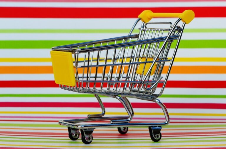A small empty shopping trolley with yellow accents sits on a multicoloured striped surface, evoking the anticipation of a fresh grocery haul or planning your next weekly meal plan against the matching striped background.