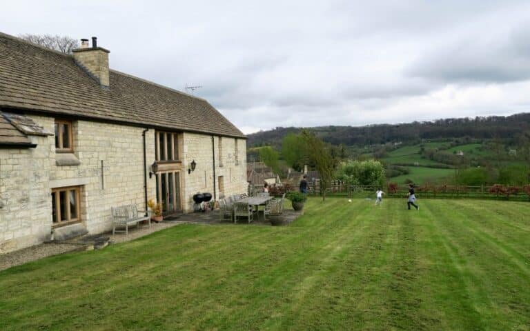 A stone house with large windows sits beside a well-kept lawn. Two children run across the grass, captured in weekend photographs, as rolling green hills and trees stretch into the distance under a cloudy sky.
