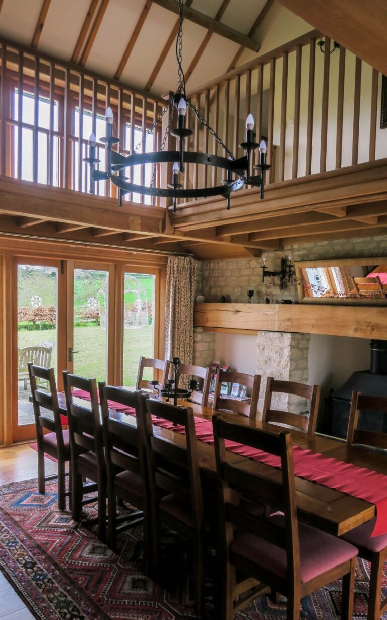 A spacious dining room at Barncastle with a long wooden table and chairs, red table runners, a large chandelier, exposed wooden beams, stone fireplace, and windows overlooking a green garden—perfect for a Cotswolds weekend away.