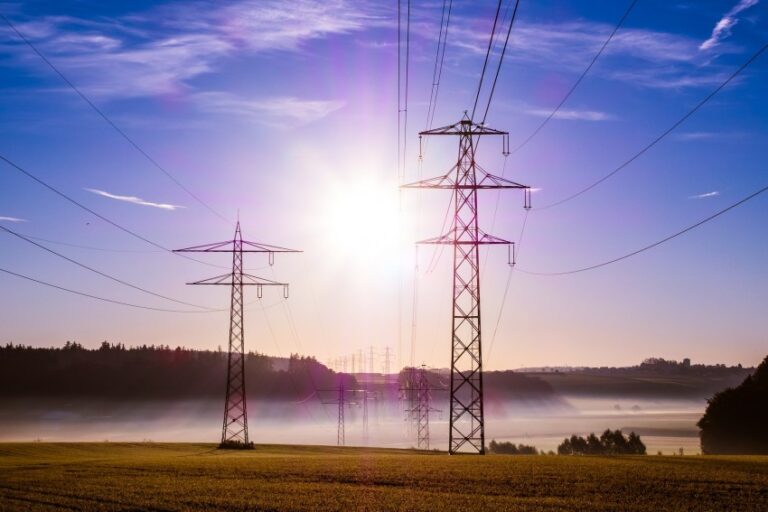 High-voltage power lines and pylons stretch across a rural landscape at sunrise, with sunlight streaming through mist over fields—an essential part of the grid that helps communities save on utilities and enjoy lower utility costs under a clear blue sky.