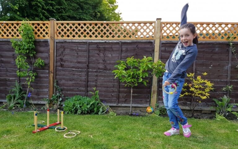 A young girl in a garden smiles excitedly with one arm raised, holding a rope ring. She appears to play a hoopla game, turning #TearsIntoSmiles as she aims for the wooden pegs set up on the grass nearby.