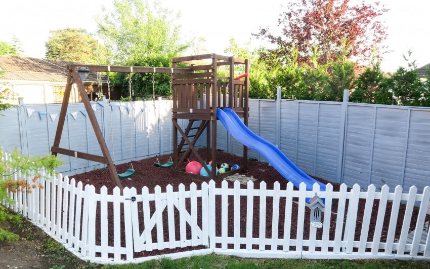 A fenced playground with a wooden climbing frame, blue slide, swings, and small toys on bark is surrounded by a white picket fence—perfect for a Garden Reveal feature or sharing inspiration on your favourite UK Home Blog.
